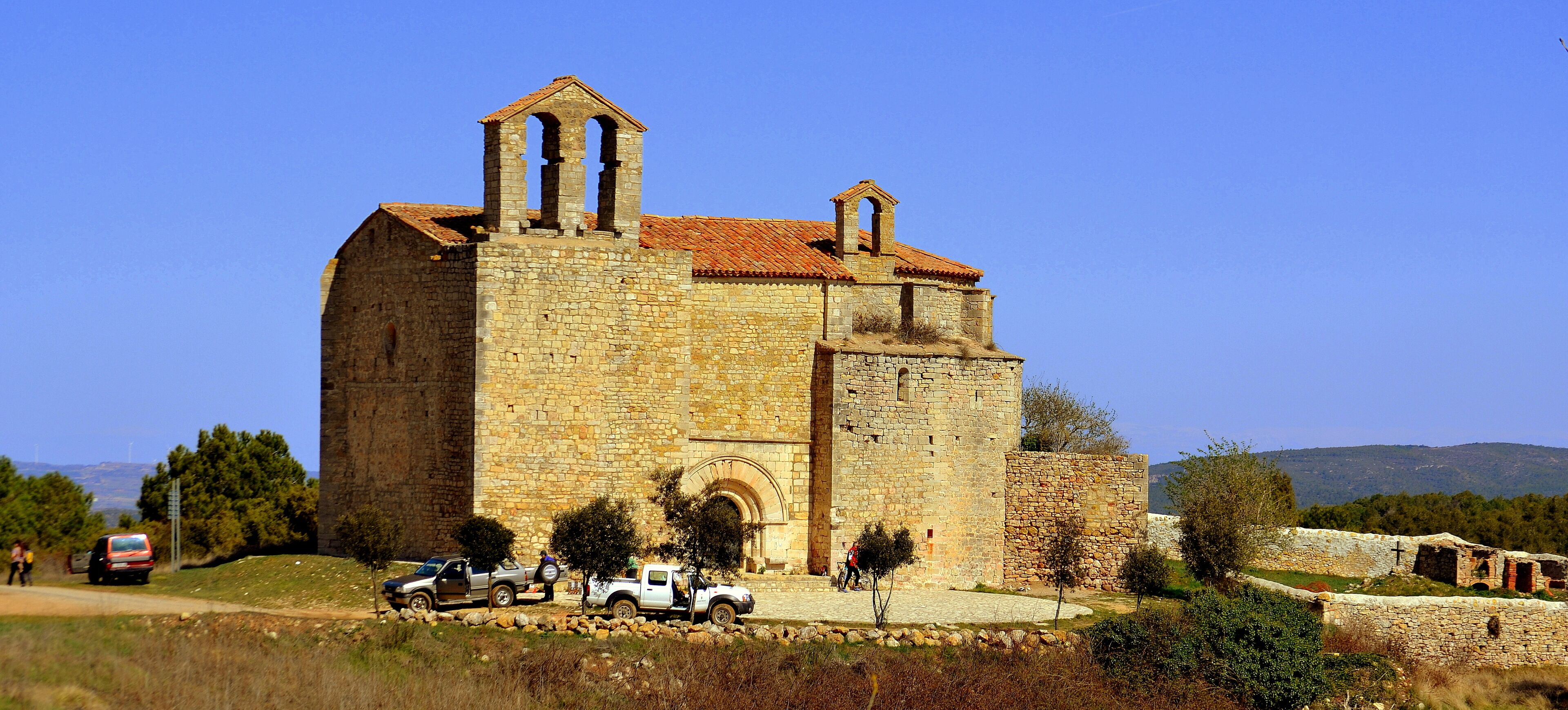 A historic stone church with a bell tower, surrounded by trees and parked vehicles.