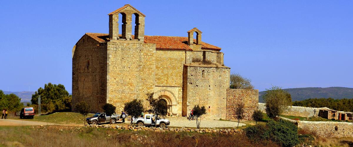 A historic stone church with a bell tower, surrounded by trees and parked vehicles.
