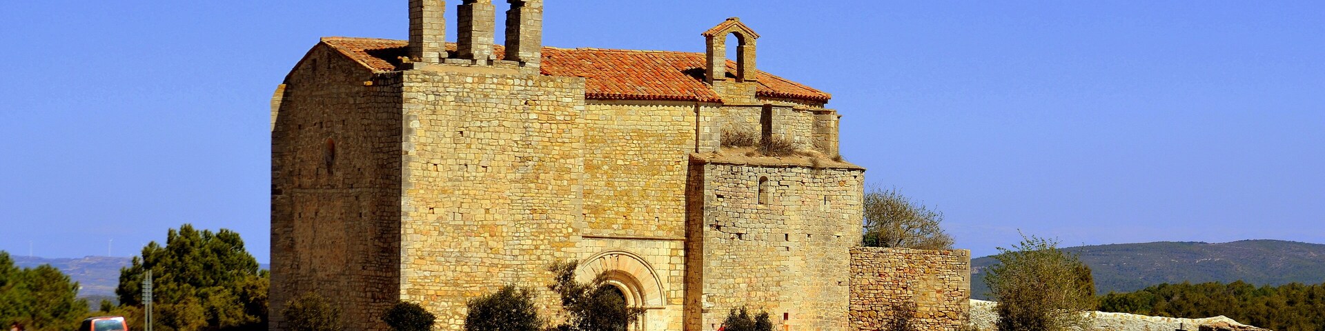 A historic stone church with a bell tower, surrounded by trees and parked vehicles.
