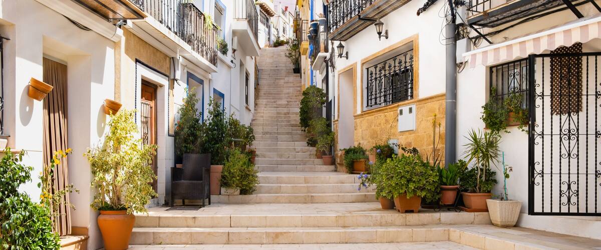 Narrow street with steps, white houses and potted plants in ancient neighborhood El Barrio or Casco Antiguo Santa Cruz in Alicante old town on hillside. Costa Blanca on Mediterranean sea coast, Spain
