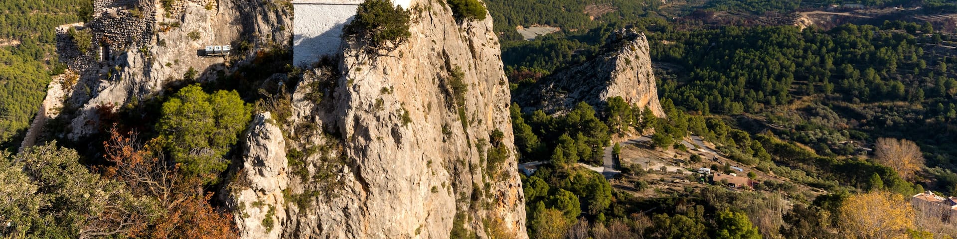 A white church with a tiled roof perched atop a rocky cliff, overlooking a valley with scattered trees and distant mountains under a clear sky.