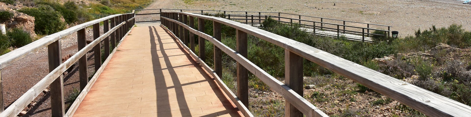 A coastal walkway leading to a sandy beach with a backdrop of residential buildings and a rocky cliff.