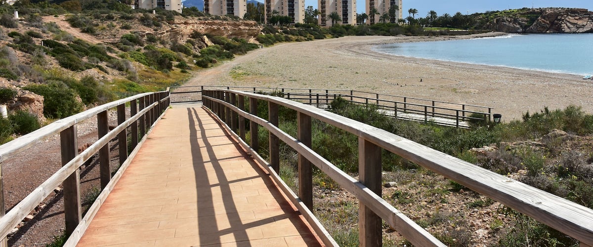 A coastal walkway leading to a sandy beach with a backdrop of residential buildings and a rocky cliff.