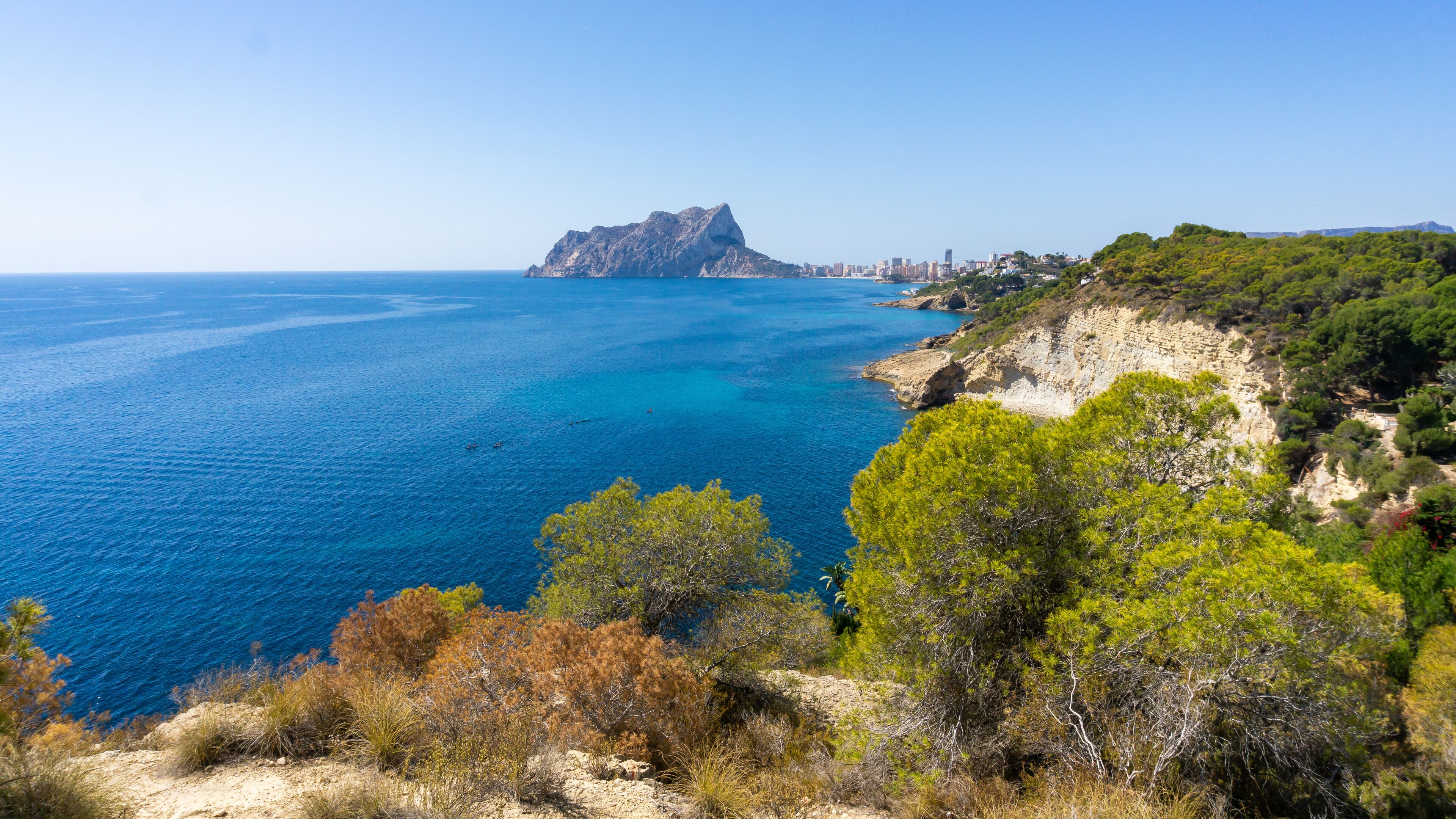 A coastal landscape with a prominent rocky outcrop, clear blue sea, and dense green vegetation.