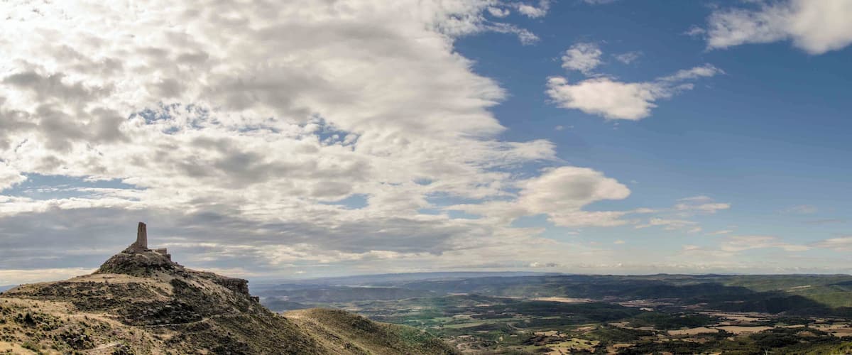 A mountain with a stone structure at its peak, a valley with scattered buildings, and a partly cloudy sky.