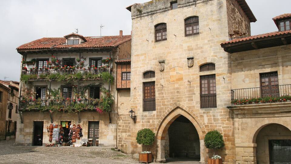 A cobblestone street with historic stone buildings, flower boxes on balconies, and a small archway entrance.
