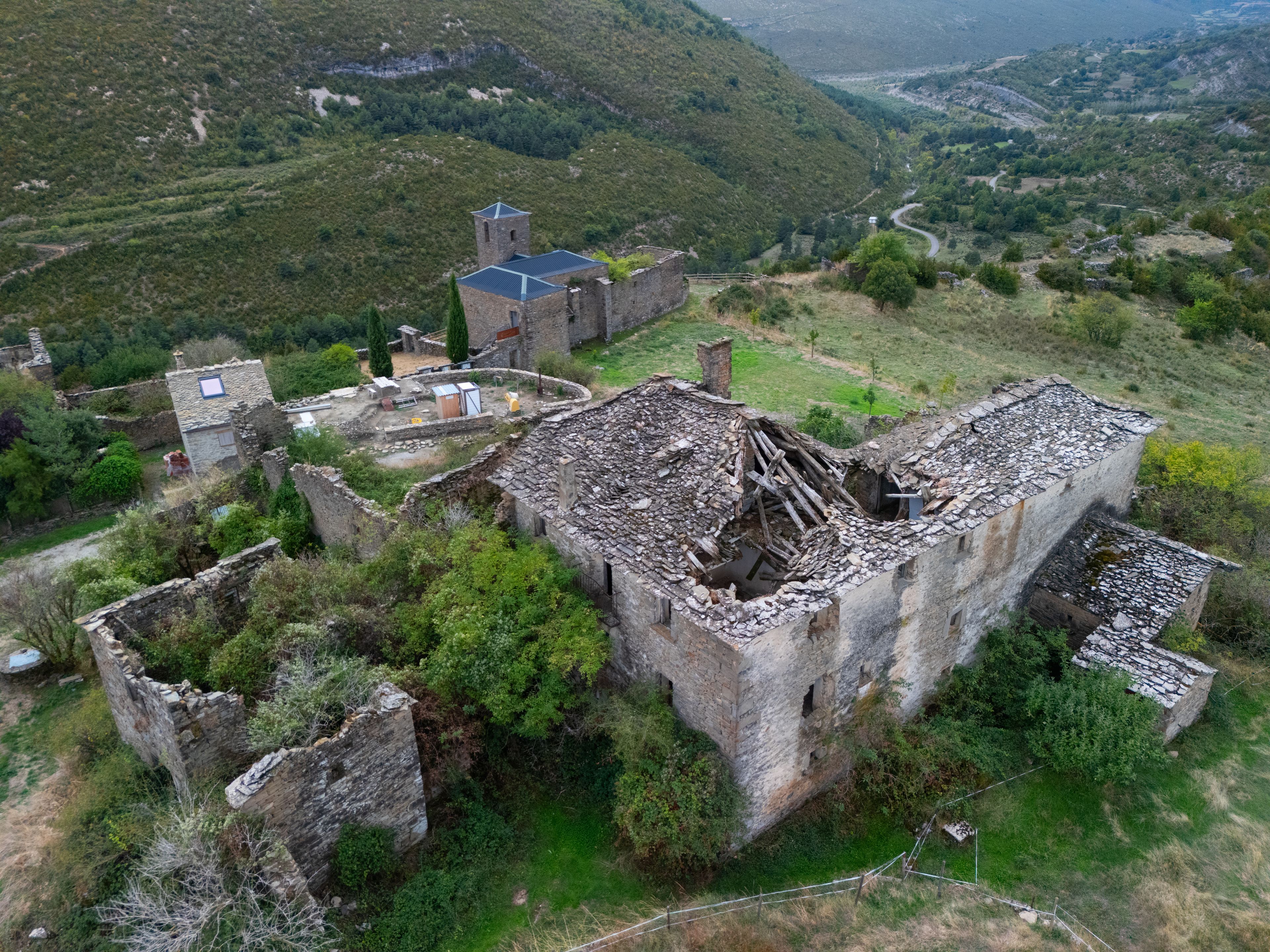 An aerial view of a historic stone building with ruins, surrounded by lush greenery and hills.