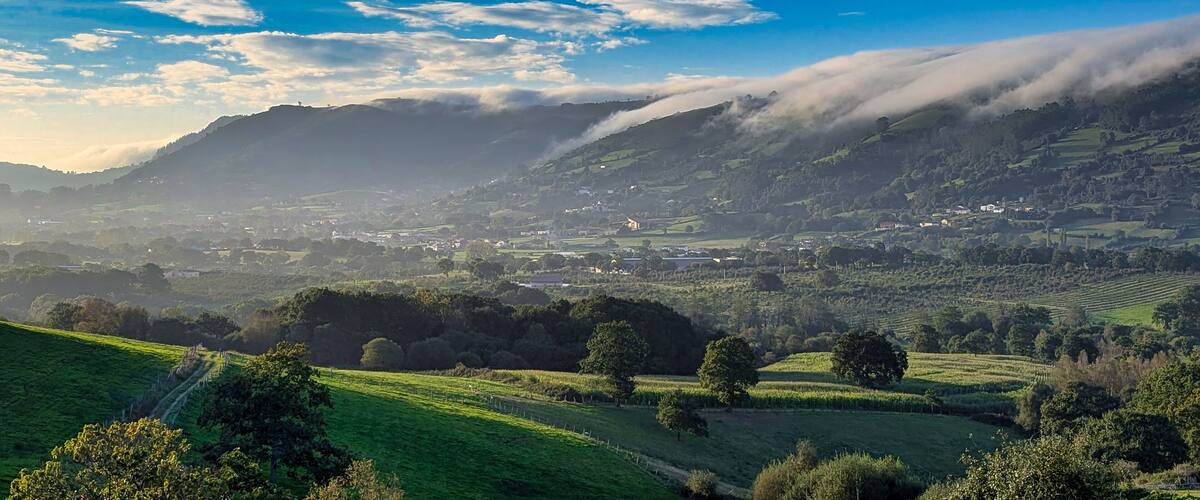 A valley with rolling hills, a river, and a mountain range with clouds.