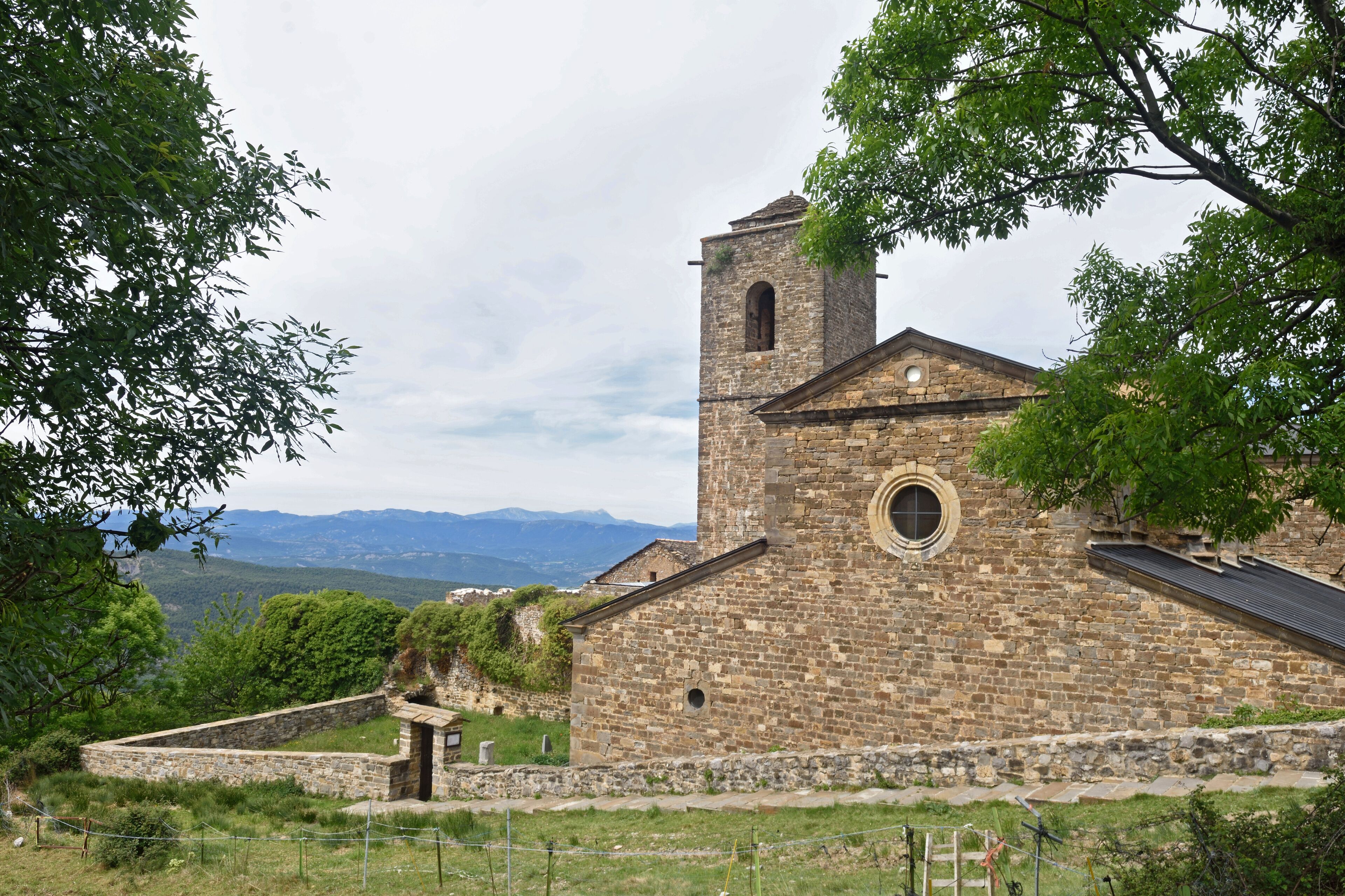 A stone church with a bell tower and a circular window, surrounded by greenery and mountains in the background.