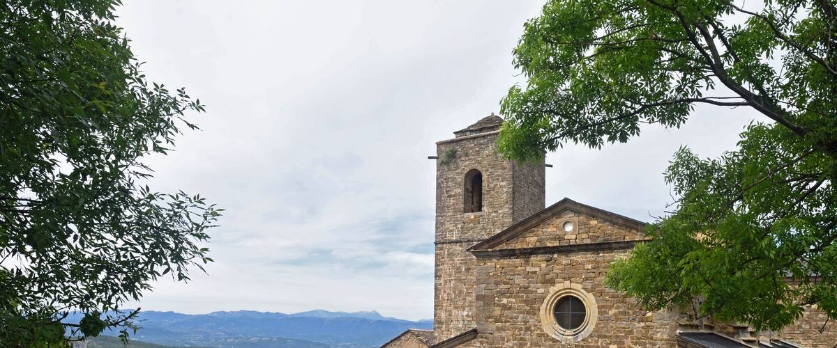 A stone church with a bell tower and a circular window, surrounded by greenery and mountains in the background.