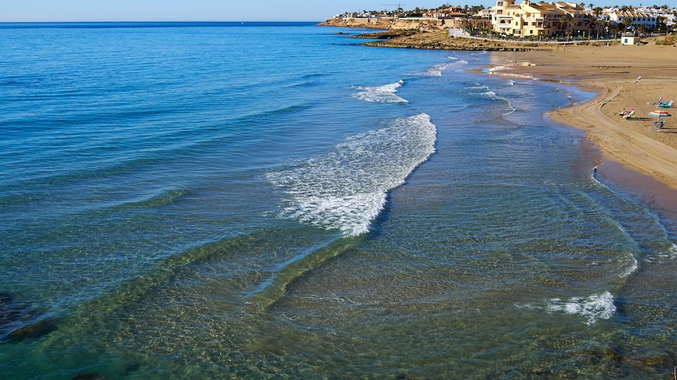 A coastal beach with clear waters, a sandy shore, and buildings in the background.