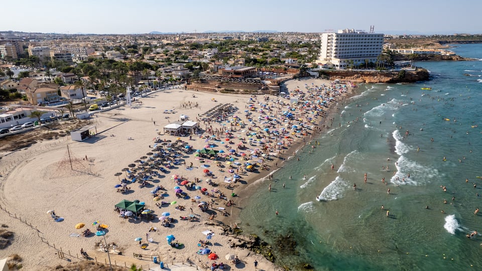 A crowded beach with numerous umbrellas and people, a large hotel, and a clear coastline.