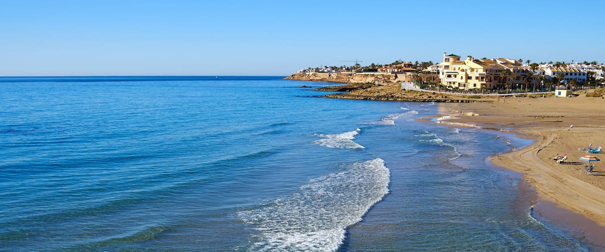 Une plage côtière avec une eau claire, des vagues et des bâtiments en arrière-plan.