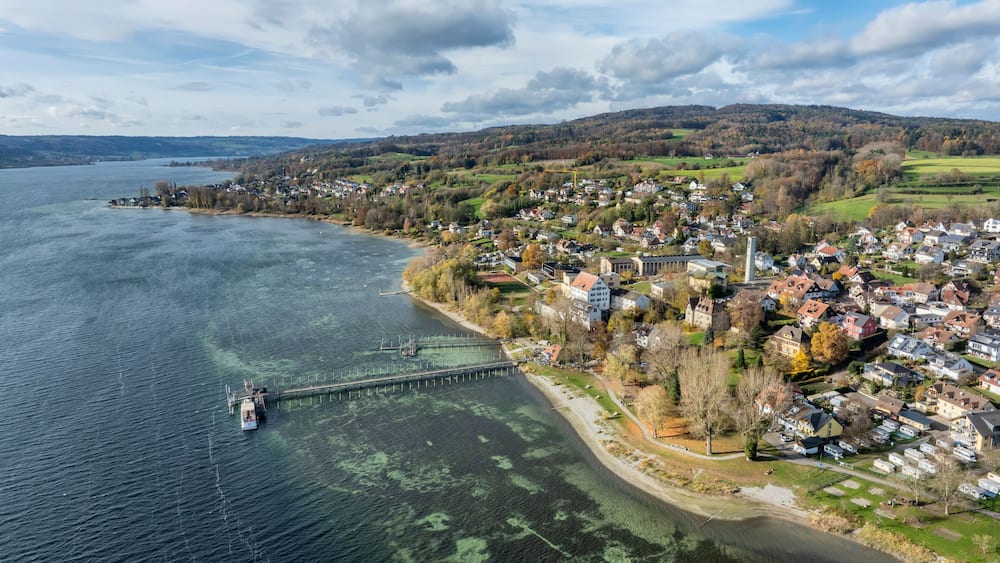 A coastal town with a pier extending into a calm lake, surrounded by hills and residential buildings.
