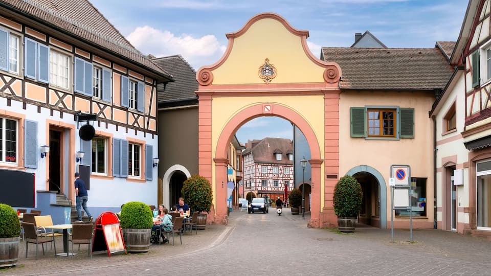 A cobblestone street with traditional half-timbered buildings, a central archway, and outdoor seating at a café.