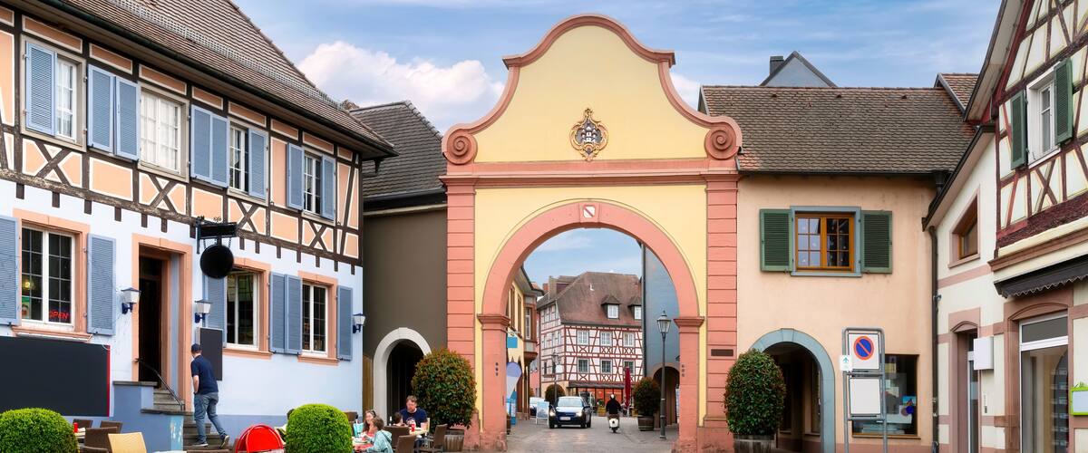 A cobblestone street with traditional half-timbered buildings, a central archway, and outdoor seating at a café.