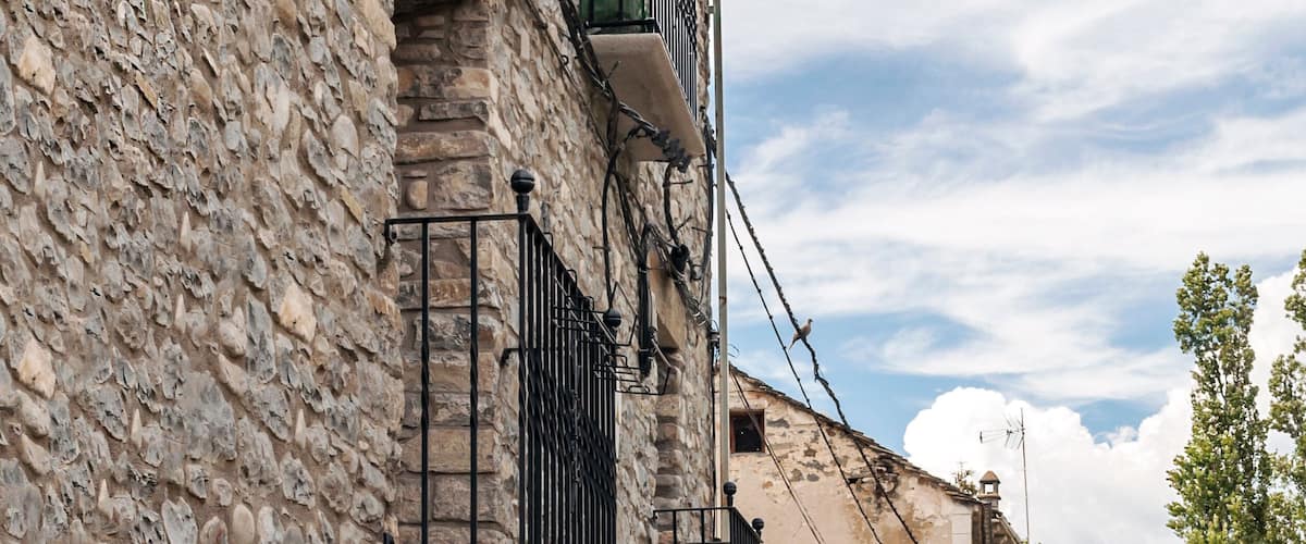 A narrow cobblestone street lined with stone buildings, featuring balconies and hanging plants.