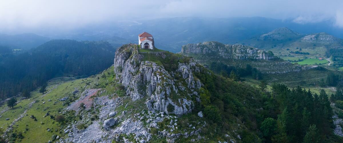 A church on a mountain peak surrounded by lush green fields and rocky terrain.
