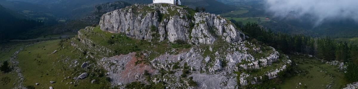 A white church on a rocky hilltop with a red roof, surrounded by a mountainous landscape and clouds.
