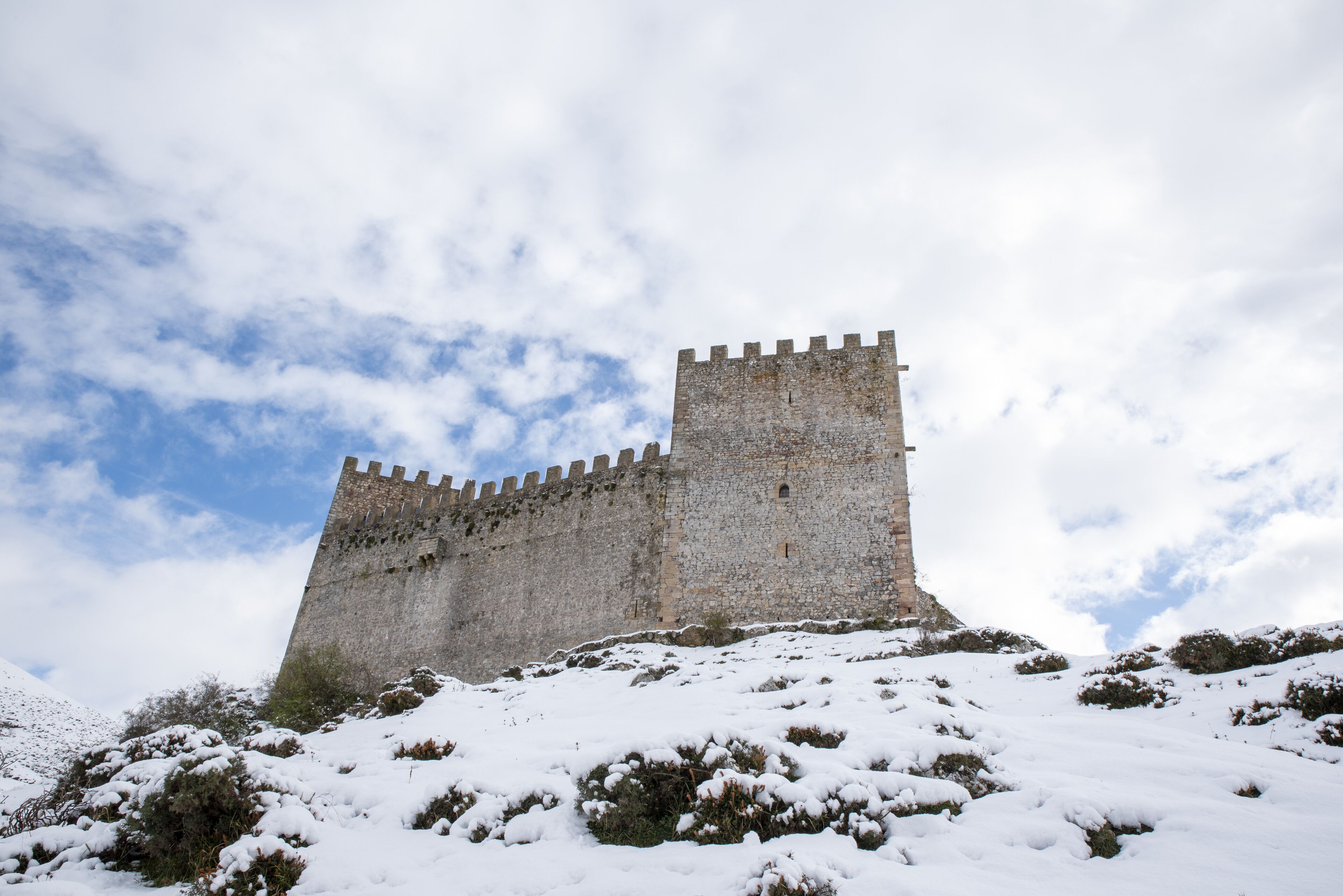 A medieval stone castle on a snow-covered hill under a partly cloudy sky.