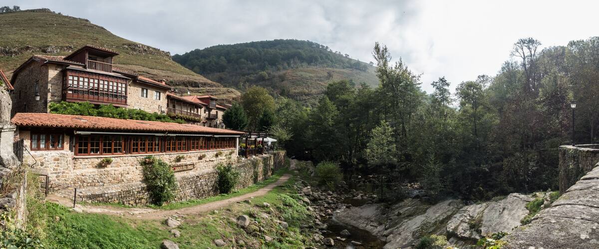 A stone building with a red-tiled roof, a stone wall, and a path leading to a small stream.