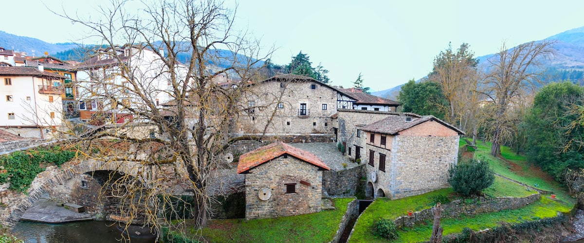 A historic stone building complex with a river, surrounded by greenery and mountains.
