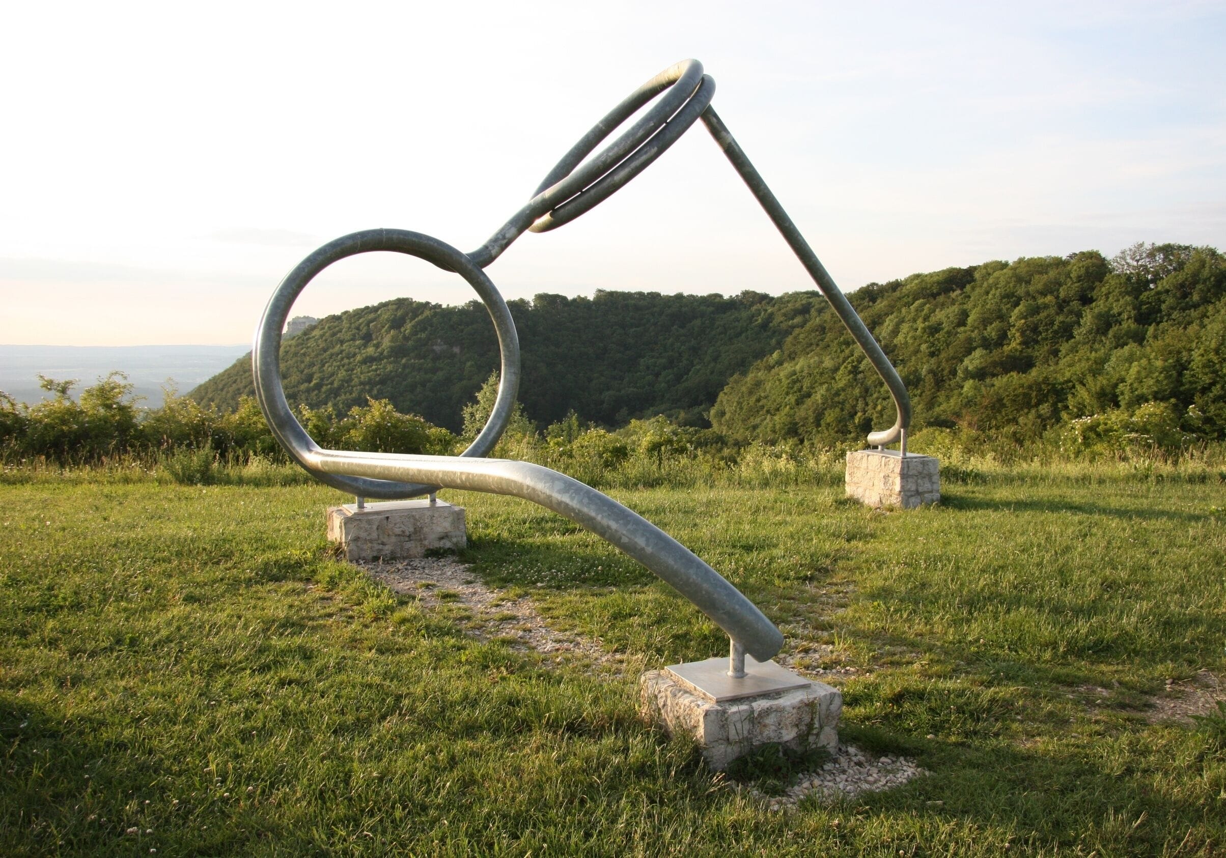 A metal sculpture in a grassy field with a forested hill in the background.