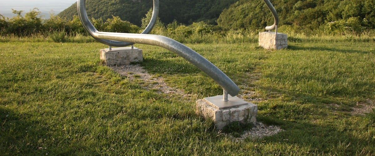 A metal sculpture in a grassy field with a forested hill in the background.