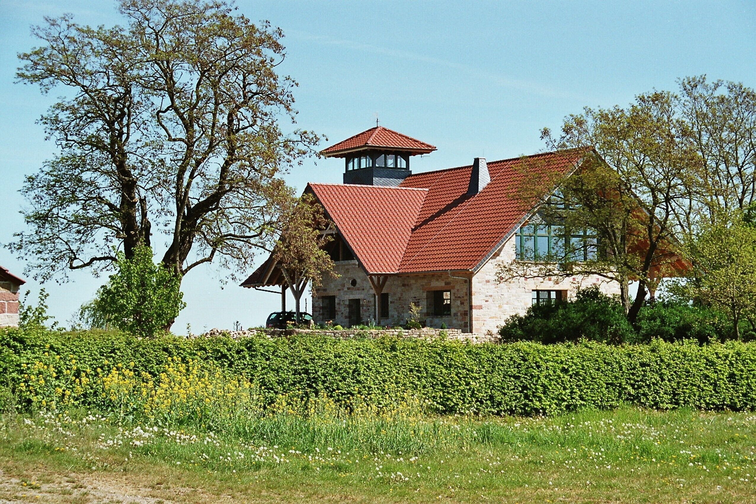 A stone house with a red roof and a tower, surrounded by trees and a field of yellow flowers.