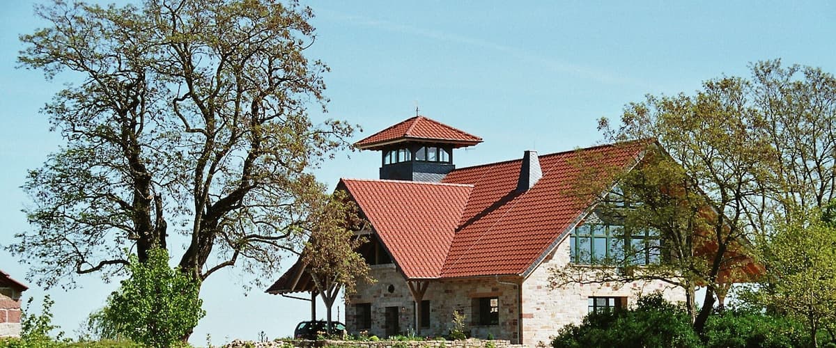 A stone house with a red roof and a tower, surrounded by trees and a field of yellow flowers.