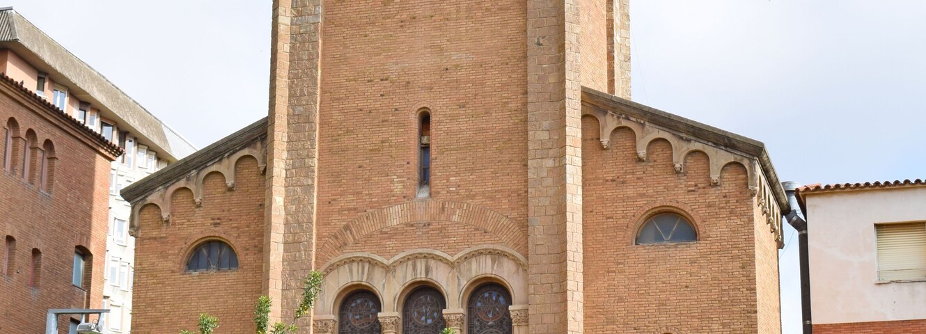 Una alta torre de ladrillo con un reloj y campanas, rodeada de árboles y bancos.