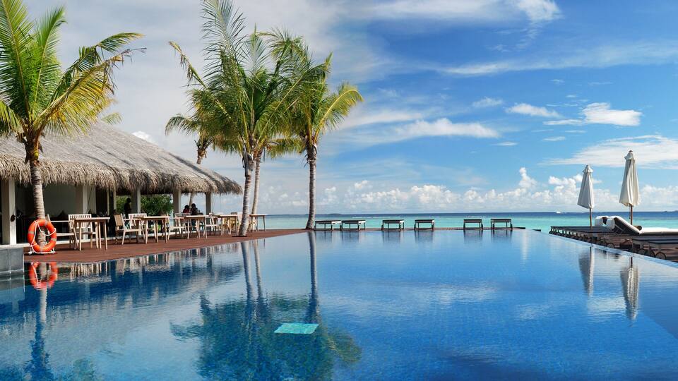 A clear blue infinity pool with lounge chairs and umbrellas, palm trees, and a thatched-roof hut by the ocean.