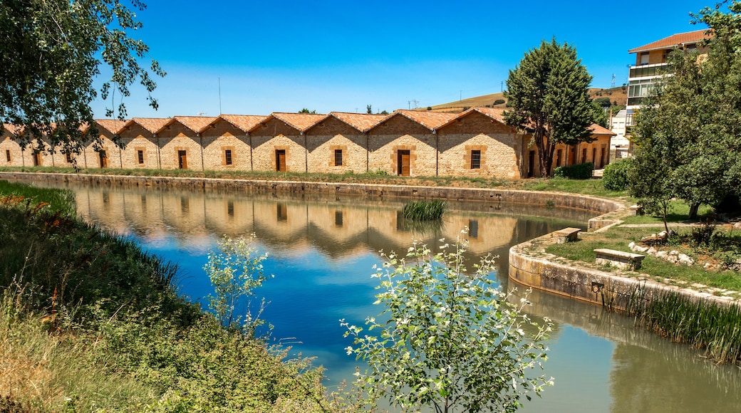 Un bâtiment en pierre surmonté d’un toit en tuiles, un plan d’eau paisible qui reflète la structure, et une végétation luxuriante tout autour.
