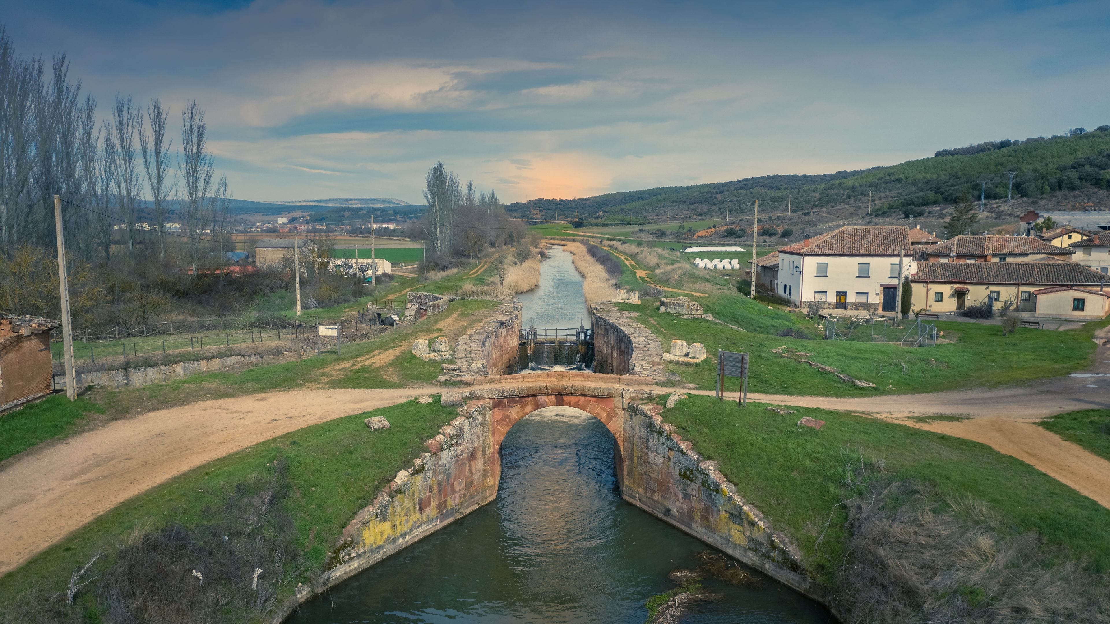 Une rivière traverse un paysage rural, avec un pont de pierre, des maisons et une colline qui se dessine à l’horizon.
