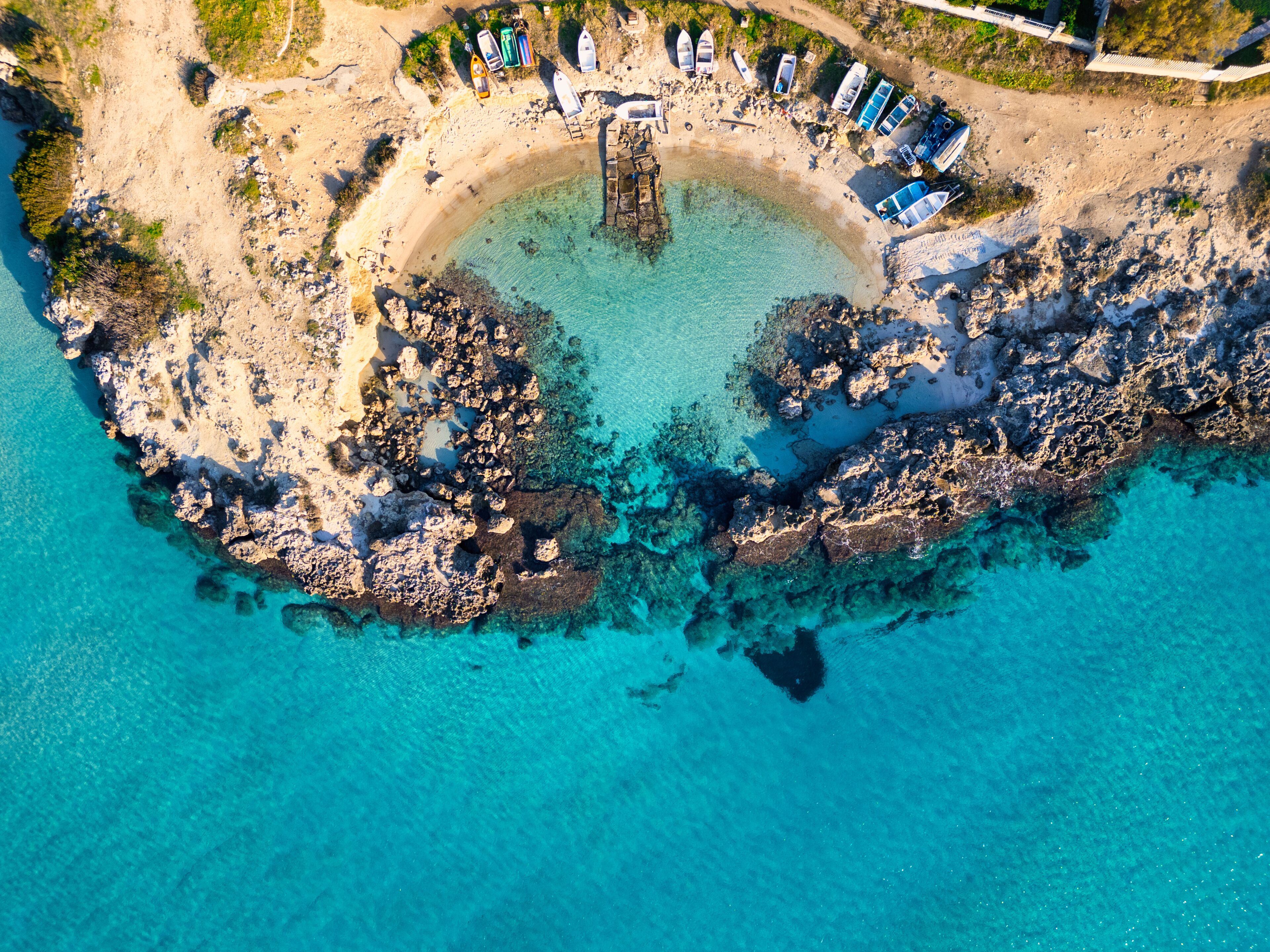 An aerial view of a rocky beach with clear turquoise water, boats, and a circular stone structure.