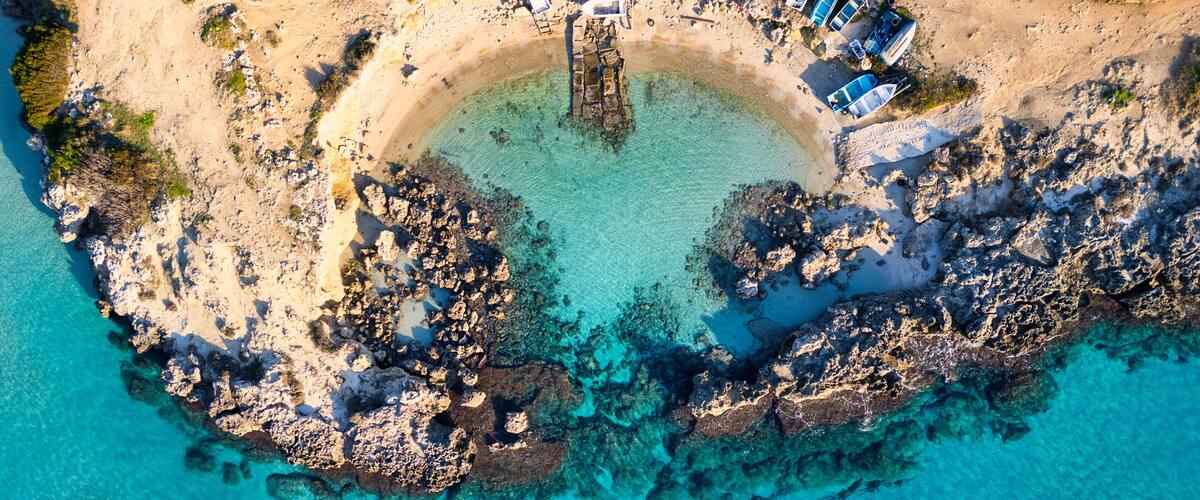An aerial view of a rocky beach with clear turquoise water, boats, and a circular stone structure.