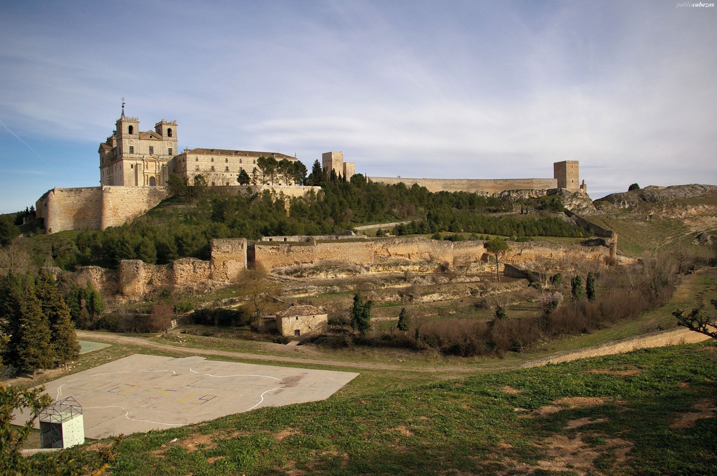 Vista del Monasterio de Uclés en la cima del cerro de El Portillo. Fue construido entre el siglo XVI y XVII sobre los restos del castillo musulmán (a la derecha del castillo). Debido al largo periodo de construcción tiene elementos platerescos, herrerianos y churrigueresco. Fue construido por la Orden de Santiago cuya casa central (Caput Ordinis) se encontraba allí. Para su construcción se utilizaron materiales de la antigua ciudad romana de Segobriga.