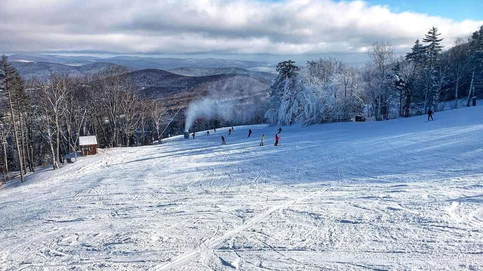 A snowy ski slope with skiers, a cabin, and a mountain range in the background.