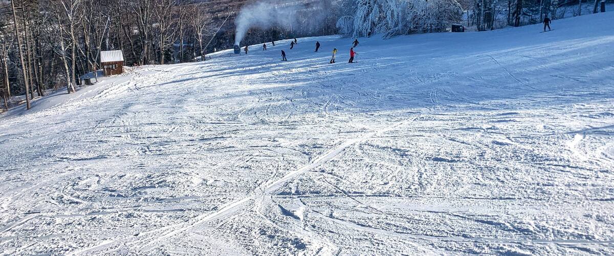A snowy ski slope with skiers, a cabin, and a mountain range in the background.