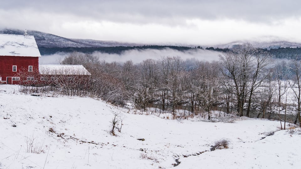 A snow-covered landscape with a red barn, leafless trees, and a valley with mist-covered mountains in the background.