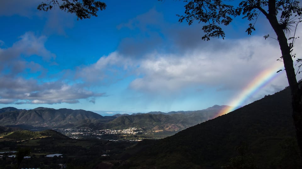 A mountain landscape with a rainbow, a town in the valley, and a tree on the right.