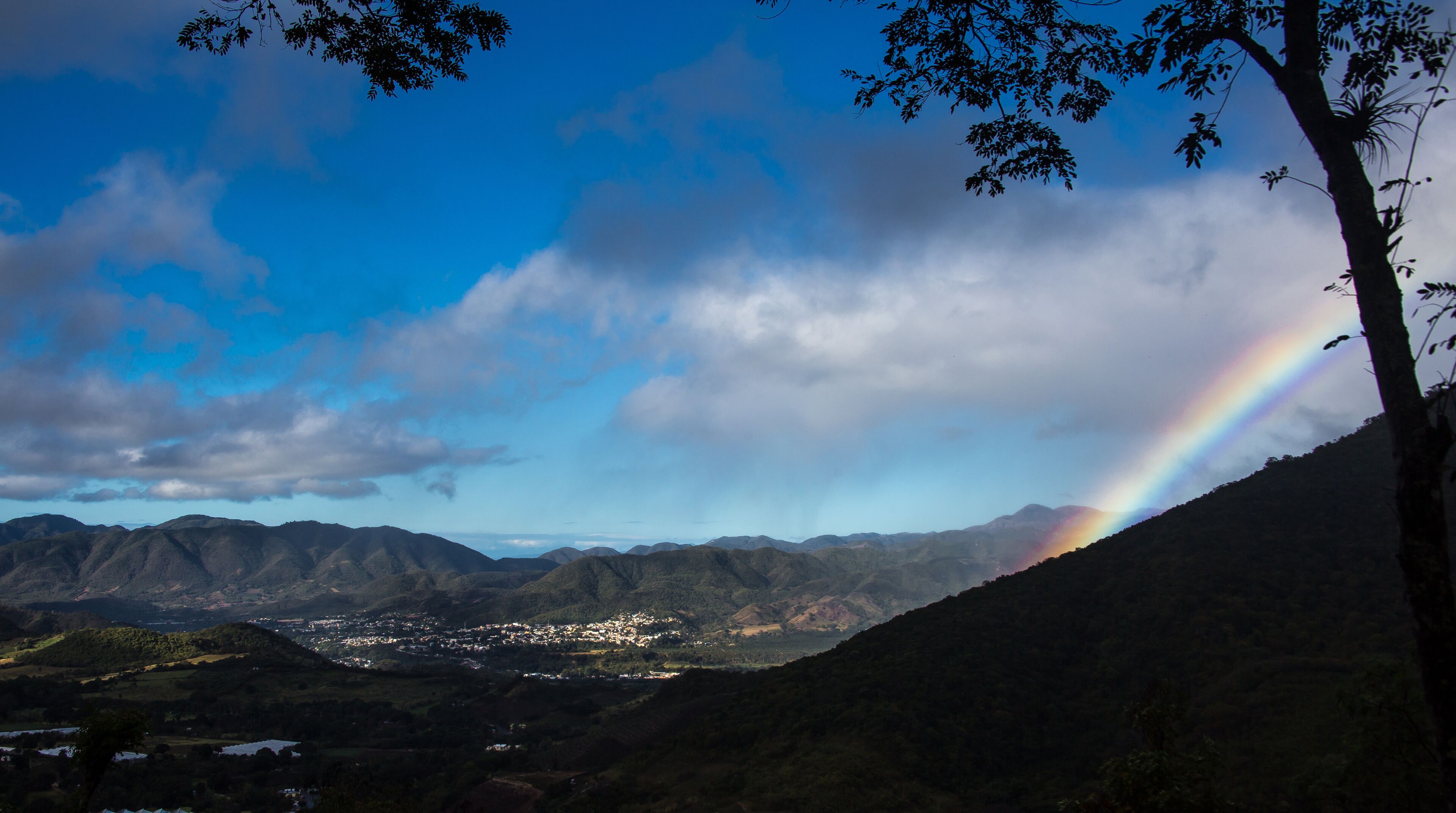 A mountain landscape with a rainbow, a town in the valley, and a tree on the right.