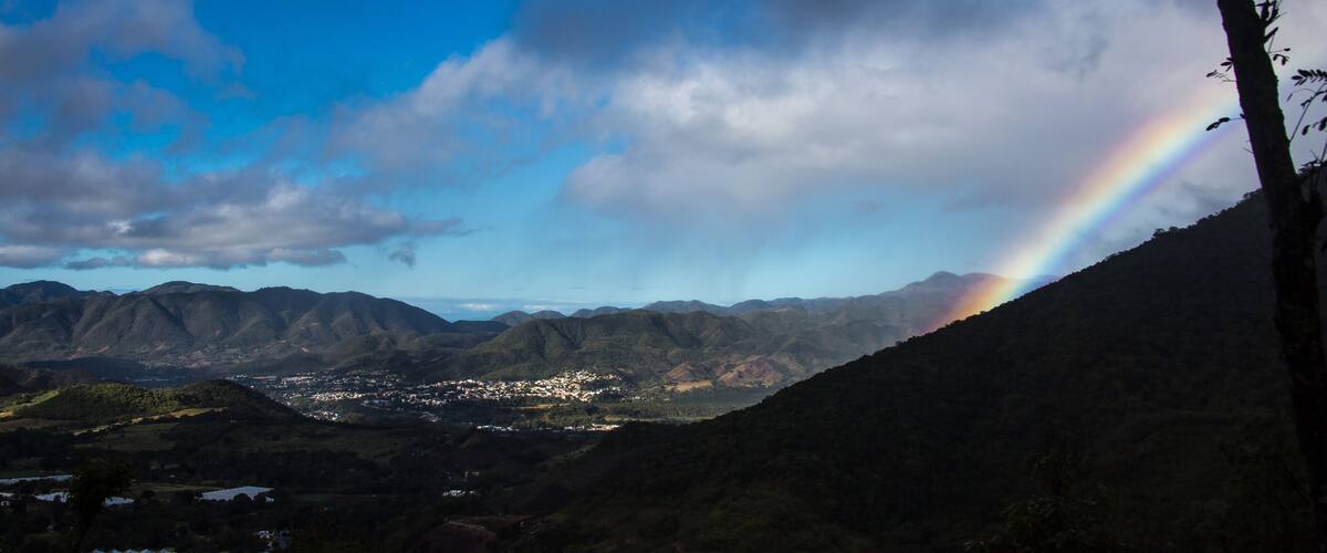 A mountain landscape with a rainbow, a town in the valley, and a tree on the right.