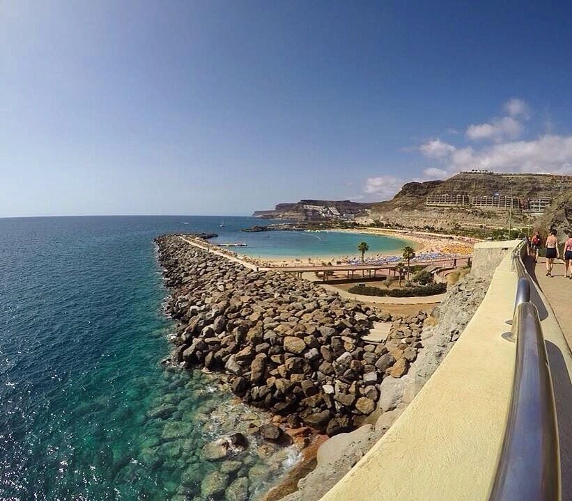 Un paseo marítimo con una barrera rocosa, aguas azul turquesa y una playa con sombrillas.