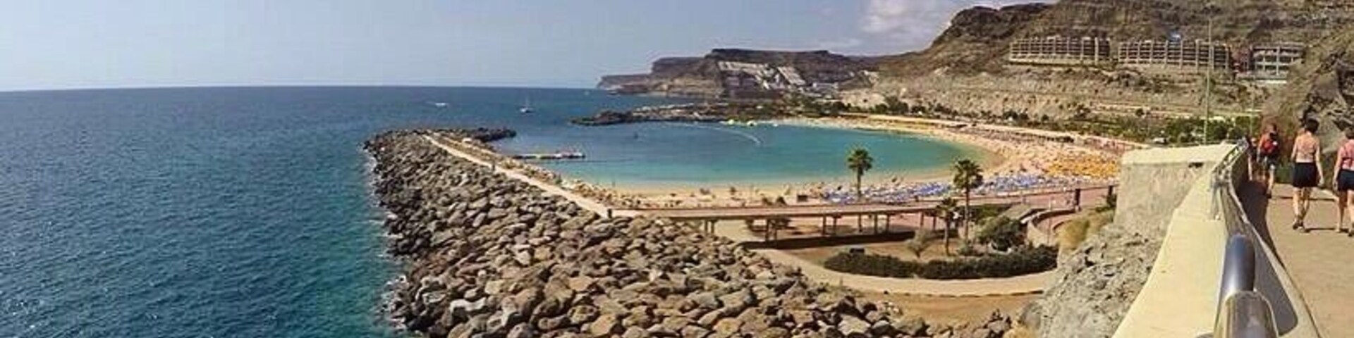 Un paseo marítimo con una barrera rocosa, aguas azul turquesa y una playa con sombrillas.