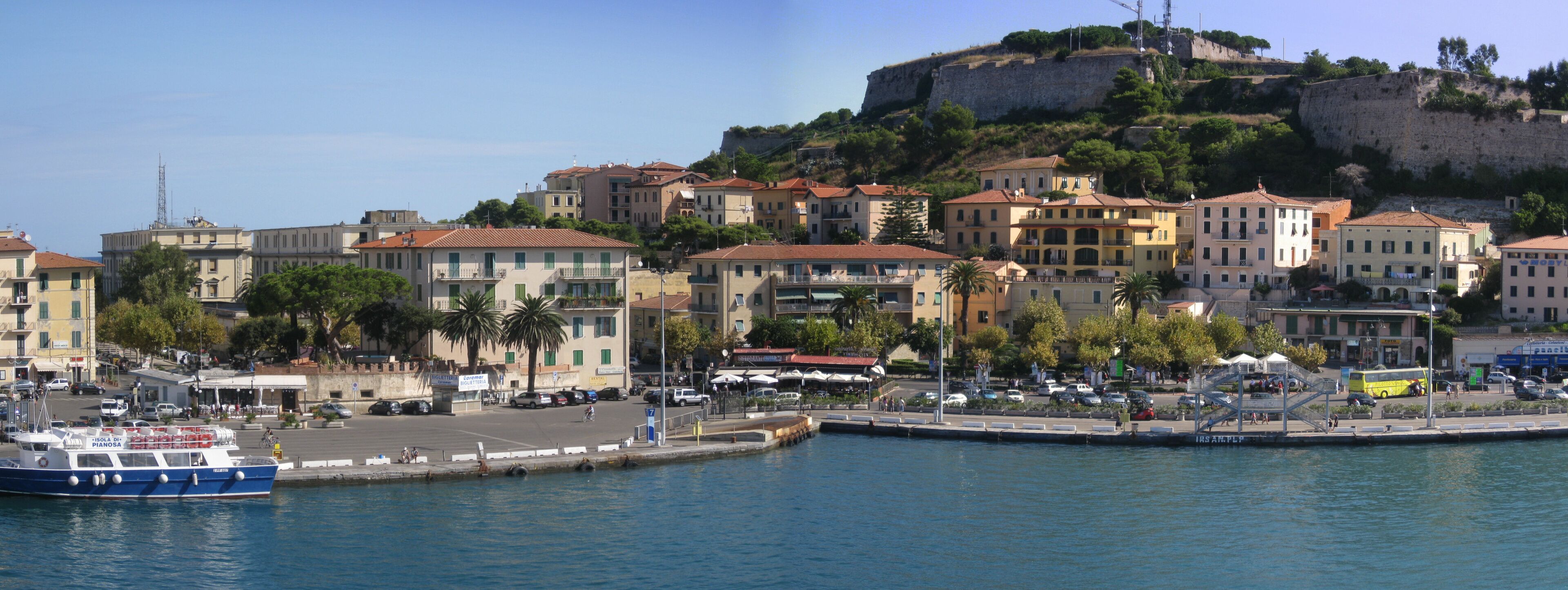 A coastal town with buildings, a dock, and a historic fort on a hill.