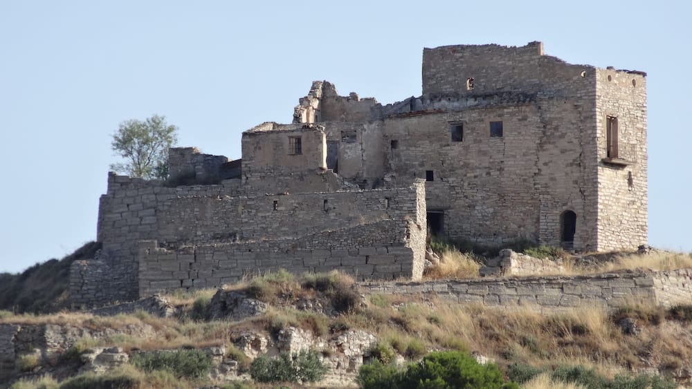 An ancient stone fortress with multiple towers and walls, surrounded by dry grass and sparse vegetation.