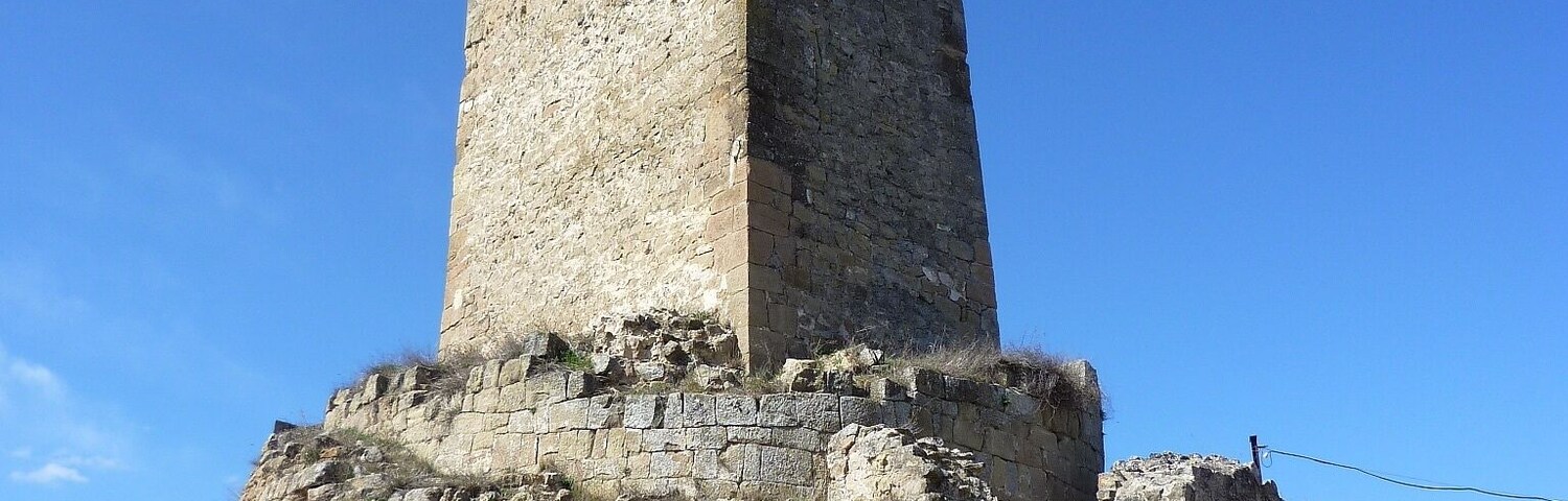 A tall, ancient stone tower with a square base, surrounded by ruins and greenery.