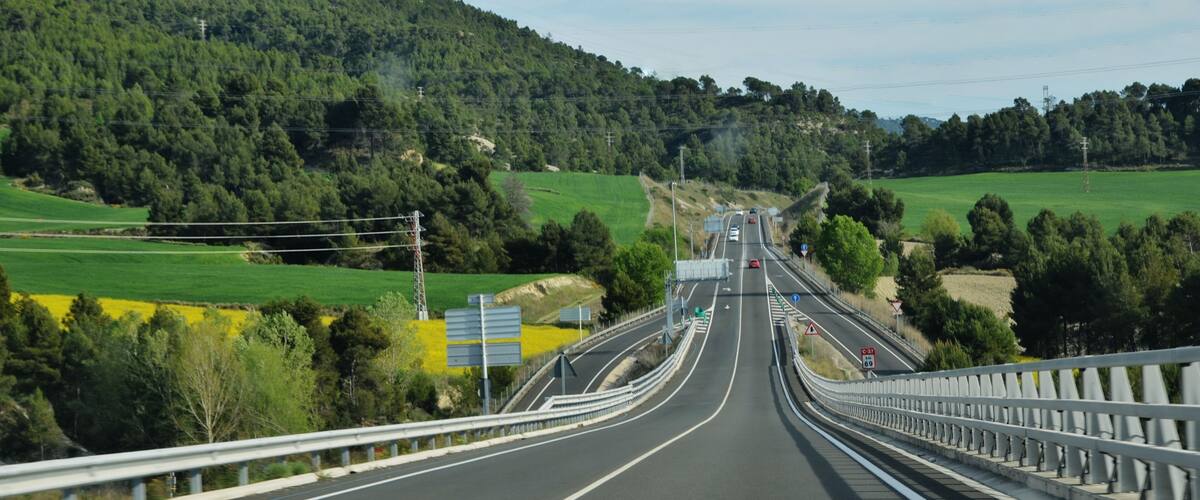 A highway with guardrails, green fields, and a forested mountain in the background.