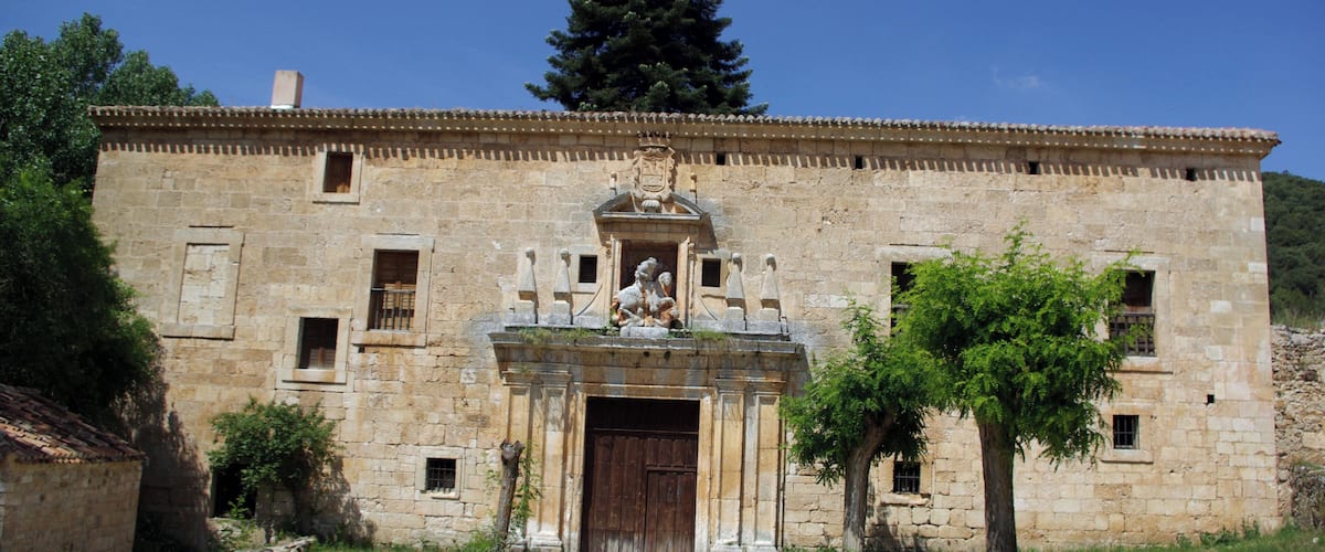 An old stone building with a wooden door, a statue above the door, and a tree in front.